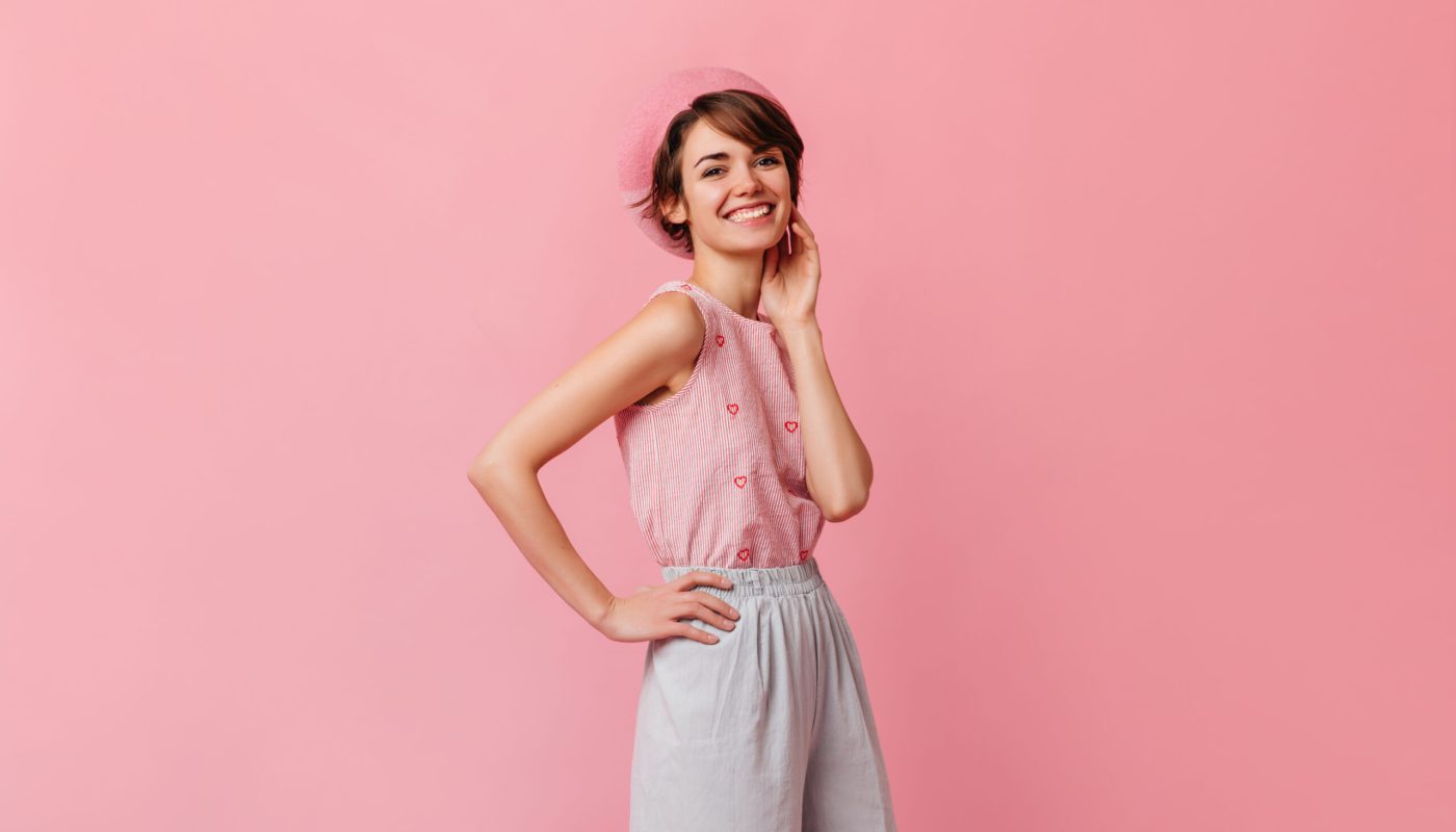 Stylish european girl in pants looking at camera. Carefree french lady standing on pink background.